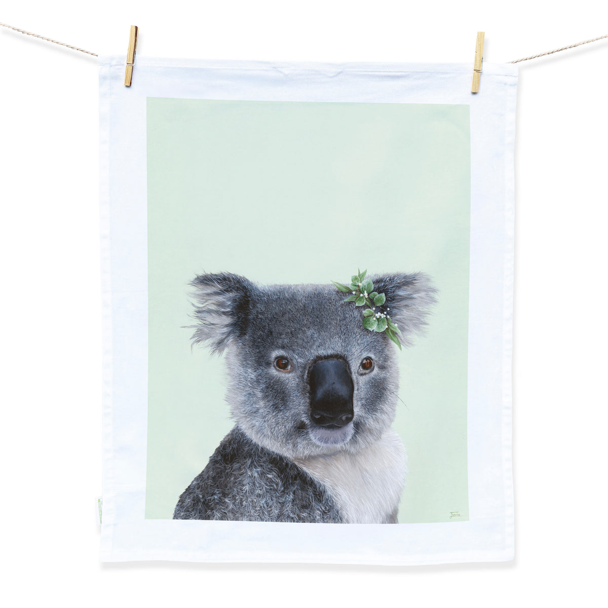 Australian Artist Jessica Alpert designed and hand painted the fluffy koala illustration. The koala is on a pale green background and has a leaf flower crown on it's right ear. The illustration is on a white tea towel with a white border hanging from two wooden pegs on a string line.