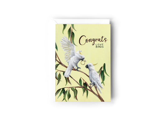 A congratulations greeting card featuring an illustration of two white sulpher crested cockatoos on a eucalyptus branch holding an engagement ring in their beak against a pale yellow background, with the text 'Congrats LOVE BIRDS' printed on the front by Australian Artist, Jessica Alpert. The card has a white envelope behind it and sits on a white background.