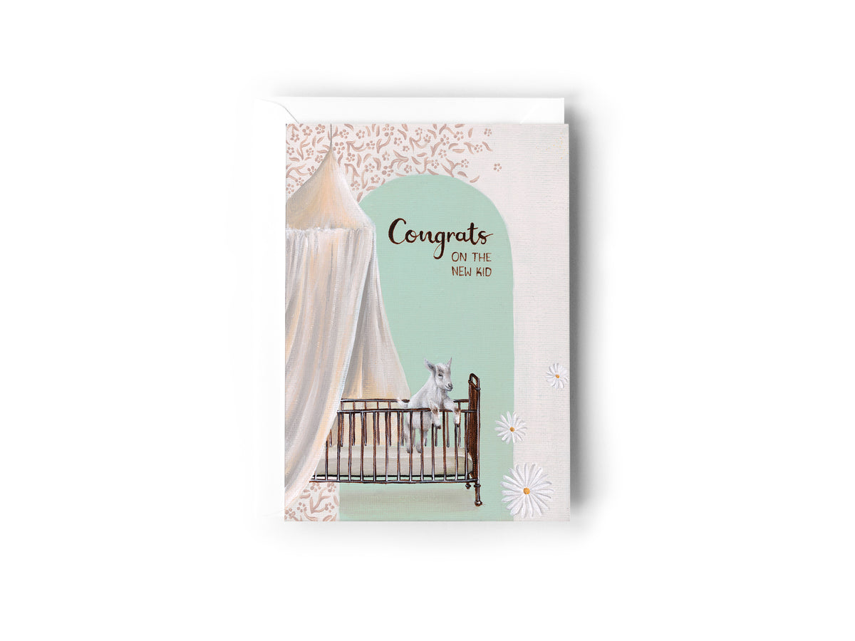 A baby shower / new baby congratulations card featuring an illustration of a white goat in a retro brass crib under a flowing bed canopy, with a mint green arch and the text 'Congrats on the NEW KID' by Australian Artist, Jessica Alpert. The card has a white envelope behind it and sits on a white background.