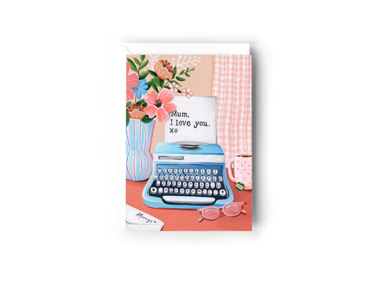 A greeting card featuring a retro illustration of a blue typewriter with a handwritten note that says 'Mum, I love you. xo', set against a pink and peach background with flowers, a mug, and pink glasses, by Australian Artist Jessica Alpert. The card has a white envelope behind it and sits on a white background.