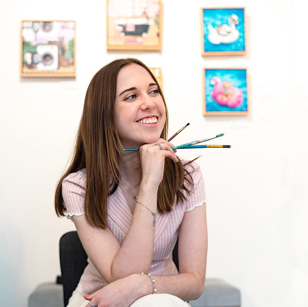 Australian Artist Jessica Alpert sitting and smiling holding paintbrushes in front of her original acrylic paintings at her 'Wild Whimsy' exhibition, held in the Yarra Valley in 2025.