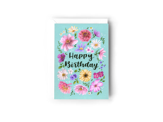 A Birthday Card design hand painted by Australian Artist Jessica Alpert featuring an array of pretty pink, purple, yellow and white flowers and green foliage against a turquoise background. The words 'Happy Birthday' are written in a script font in the centre of the card surrounded by the florals and white dots. The card has a white envelope behind it and sits on a white background.