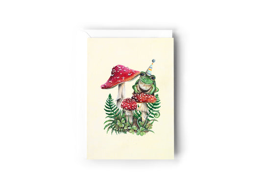 A Birthday Card design hand painted by Australian Artist Jessica Alpert of a green frog in a party hat sitting on a red mushroom with white spots in amongst other mushrooms and green foliage. The frog is smiling and wearing a striped party hat. The card has a white envelope behind it and sits on a white background.