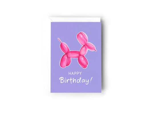A birthday card design hand painted by Australian Artist Jessica Alpert featuring a pink balloon dog illustration with a white outline on a purple background. White text saying 'Happy Birthday!' is below the balloon dog. The card has a white envelope behind it and sits on a white background.