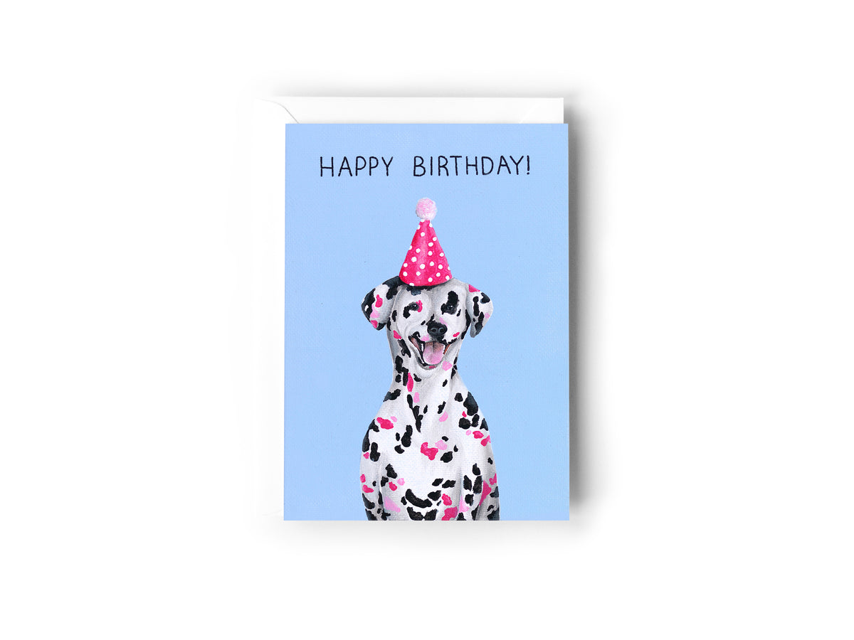 A birthday card design hand painted by Australian Artist Jessica Alpert of a smiling dalmatian dog with black and pink spots wearing a pink polka dot party hat. The card has a blue background and has the words 'Happy Birthday' written at the top.