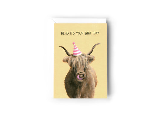 A Birthday Card design hand painted by Australian Artist Jessica Alpert featuring an illustration of a fluffy highland cow wearing a pink and white striped party hat and licking it's nose. The background is a soft warm yellow with the text 'Herd It's Your Birthday' written at the top of the card. The card has a white envelope behind it and sits on a white background.