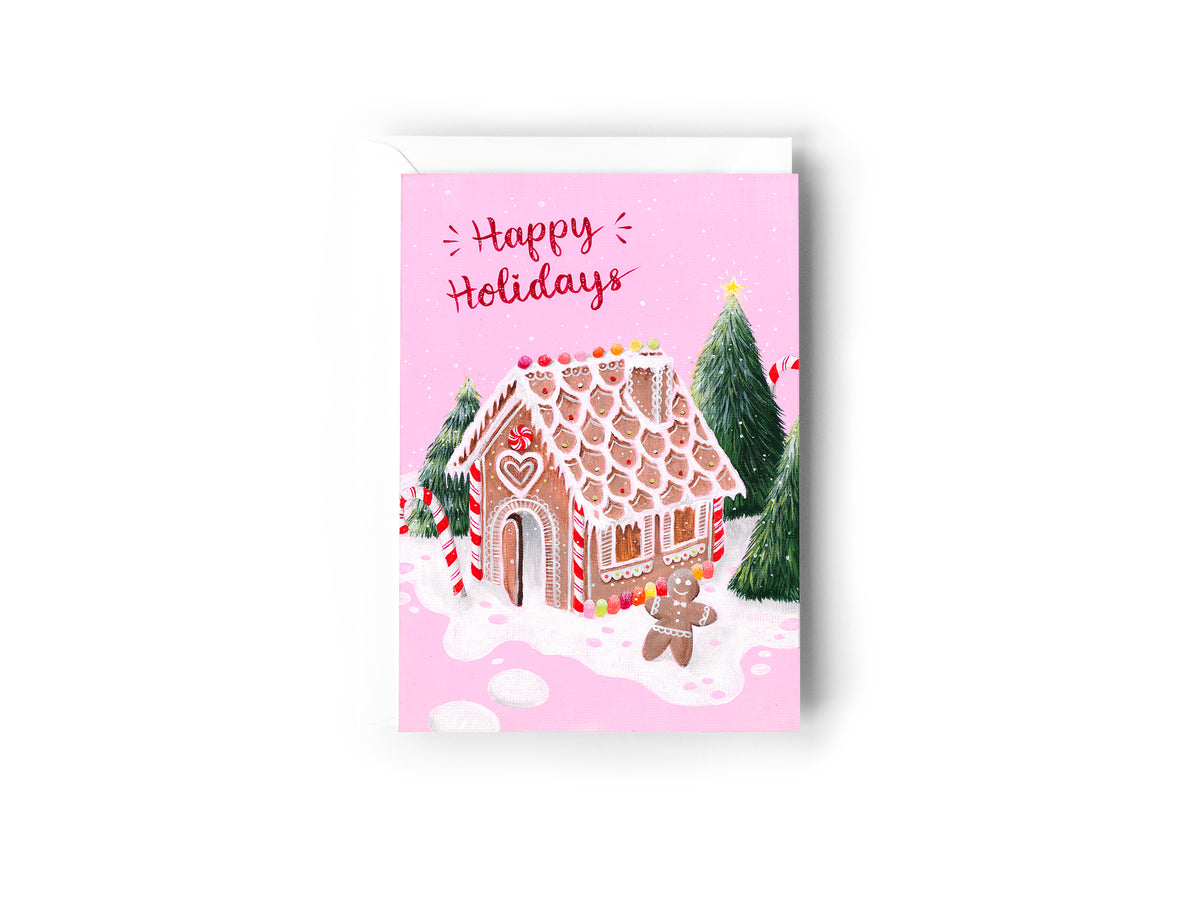 A Christmas Card design hand painted by Australian Artist Jessica Alpert of a lolly decorated gingerbread house and gingerbread man, set on a bed of icing with green Christmas trees surrounding it. Sparkly white stars and snow cover the pastel pink background, with the words 'Happy Holidays' written in a red sparkly font at the top of the card. The card has a white envelope behind it and sits on a white background.