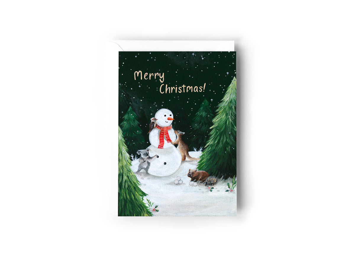 A Christmas card design hand painted by Australian Artist Jessica Alpert, of a snowy nigh time scene set amongst green Christmas trees, with a snowman being created by an Australian Kangaroo, koala, Wombat, Echidna and Kookaburra. The words 'Merry Christmas' are written in the star and snow filled sky. The card has a white envelope behind it and sits on a white background.