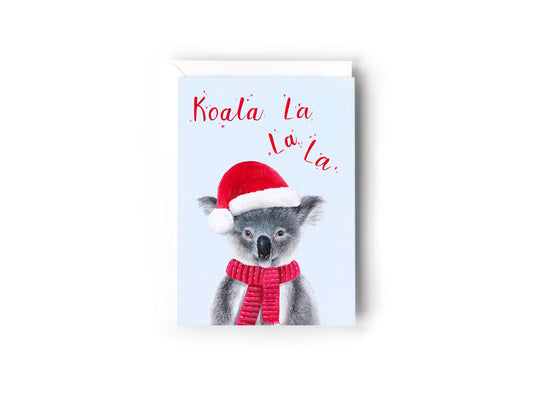 A Christmas card design hand painted by Australian Artist Jessica Alpert of a fluffy koala wearing a red Christmas hat against a pale icy blue background. The words 'Koala La La La' are written in sparkly red above the koala. The card has a white envelope behind it and sits on a white background.