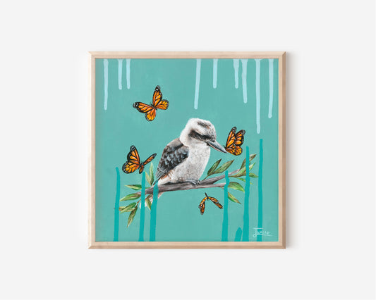 A Fine Art Print hand painted by Australian Artist Jessica Alpert of a peaceful Kookaburra perched on a branch with green leaves against a vibrant turquoise background. Pale blue and deep turquoise paint drips extend from the top and bottom of the canvas. 4 orange monarch butterflies surround the Kookaburra, with one landing on it's beak.