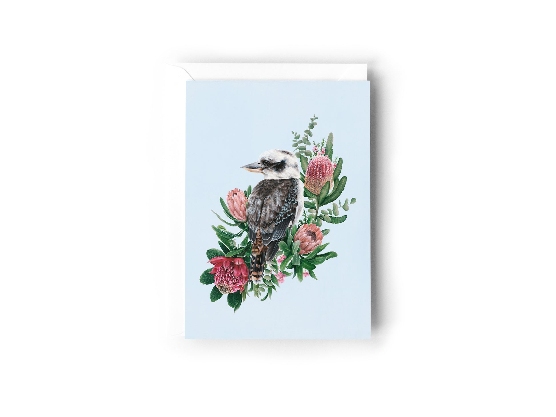 A greeting card design hand painted by Australian Artist Jessica Alpert of a peaceful kookaburra sitting against an array of green and pink native flowers and foliage, looking out against a soft blue background. The card has a white envelope behind it and sits on a white background.