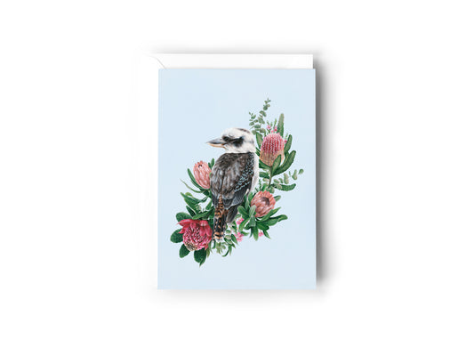 A greeting card design hand painted by Australian Artist Jessica Alpert of a peaceful kookaburra sitting against an array of green and pink native flowers and foliage, looking out against a soft blue background. The card has a white envelope behind it and sits on a white background.