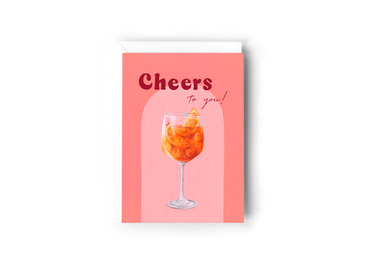 A greeting card design hand painted by Australian Artist Jessica Alpert, of an Aperol Spritz glass with a pink arch in the background against a peach solid background. The words 'Cheers to you!' are written above the glass. The card has a white envelope behind it and sits on a white background.