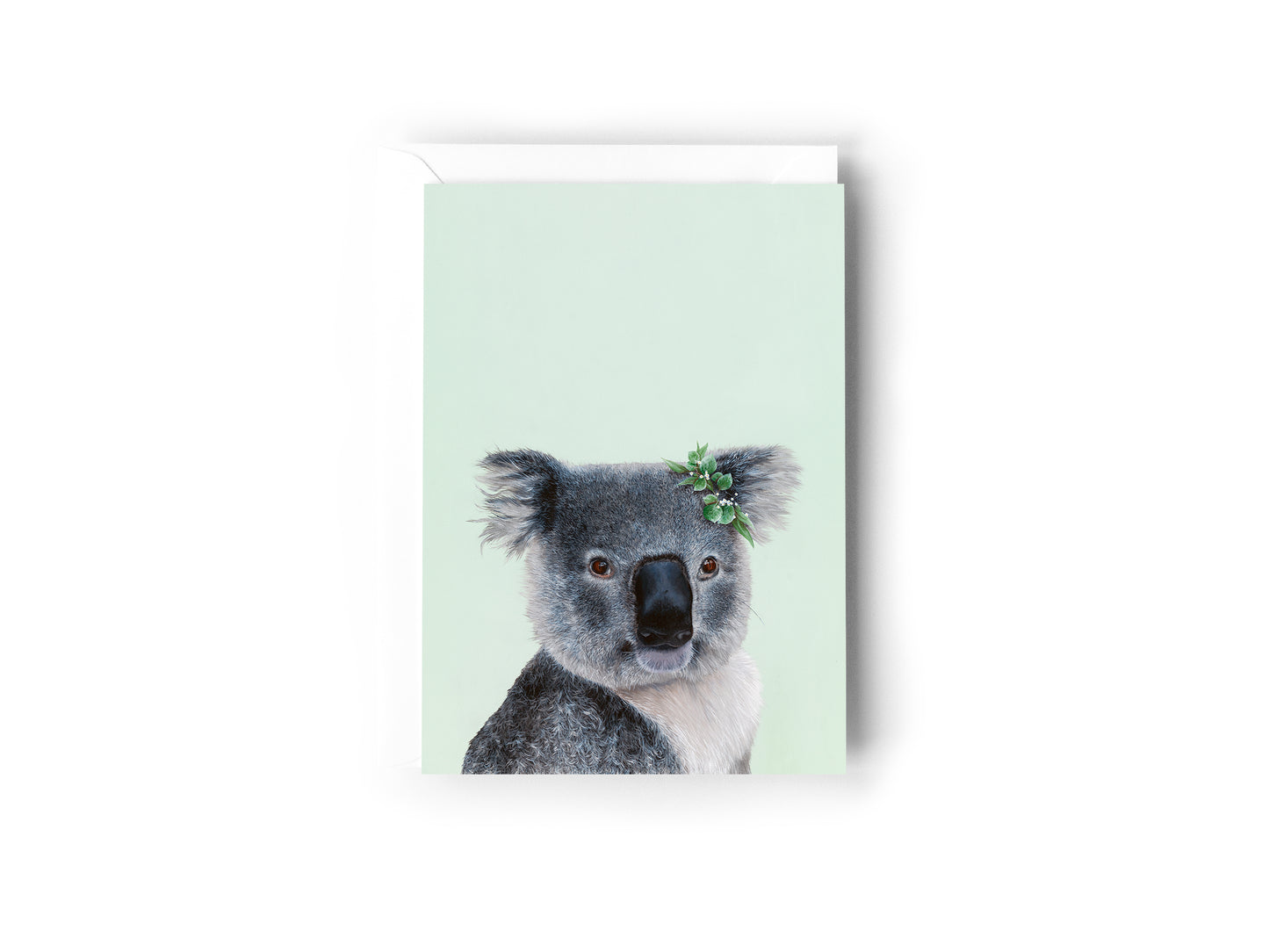 A greeting card design hand painted by Australian Artist Jessica Alpert of a detailed fluffy koala with a leaf crown on it's right ear, smiling and sitting against a soft green background. The card has a white envelope behind it and sits on a white background.
