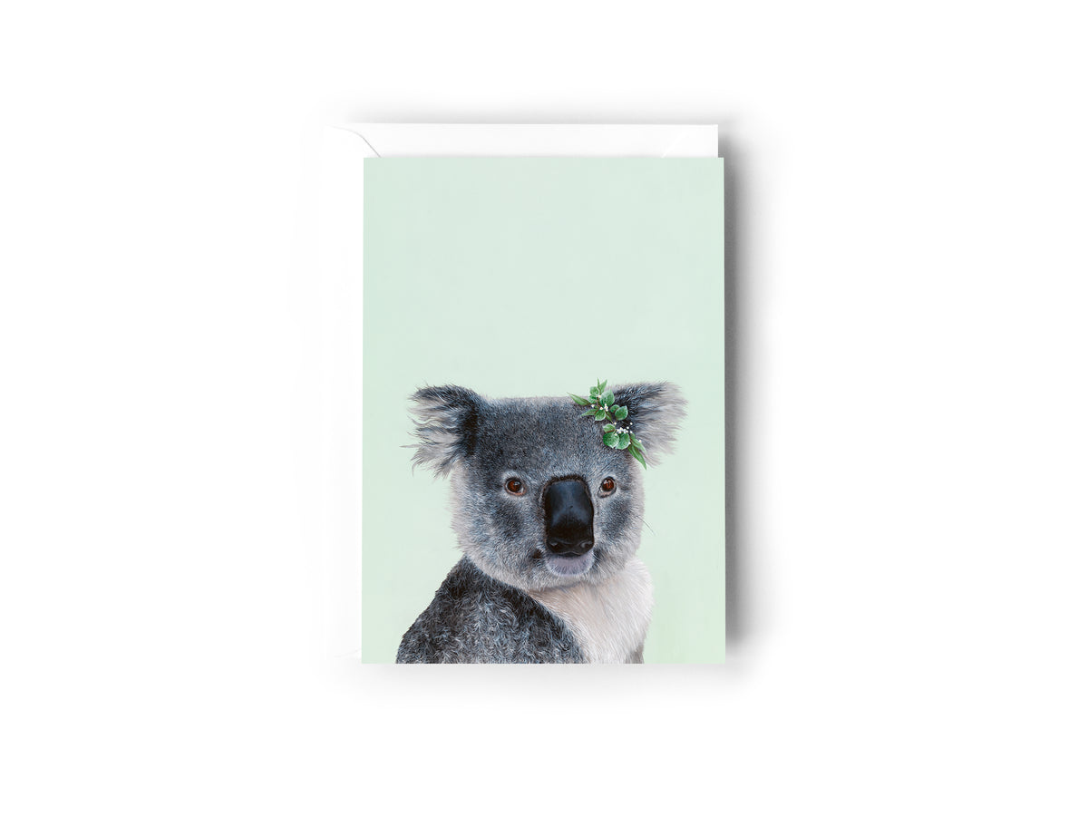 A greeting card design hand painted by Australian Artist Jessica Alpert of a detailed fluffy koala with a leaf crown on it's right ear, smiling and sitting against a soft green background. The card has a white envelope behind it and sits on a white background.