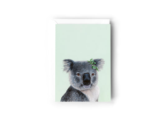 A greeting card design hand painted by Australian Artist Jessica Alpert of a detailed fluffy koala with a leaf crown on it's right ear, smiling and sitting against a soft green background. The card has a white envelope behind it and sits on a white background.