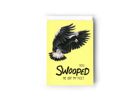 A greeting card design hand painted by Australian Artist Jessica Alpert, with the text 'You Swooped Me Off My Feet' and an illustration of a black and white Australian magpie, in flight in swooping position. The card is on a vibrant yellow background. The card has a white envelope behind it and sits on a white background.