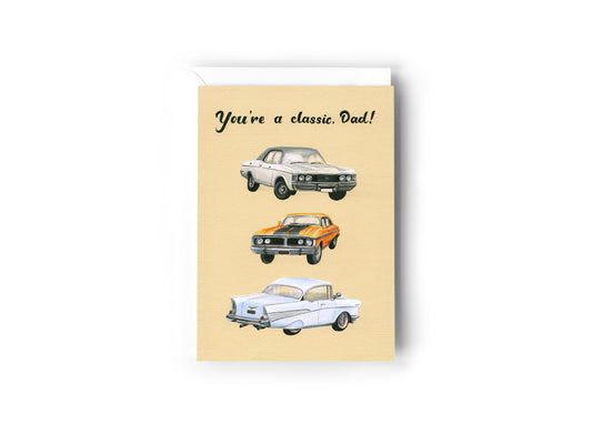 A Greeting Card design hand painted by Australian Artist Jessica Alpert of three classic cars aligned vertically on a cream background. The text, 'You're A Classic Dad!' is written in a script font at the top of the card. The card has a white envelope behind it and sits on a white background.