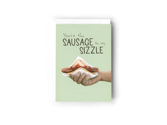 A Greeting Card design hand painted by Australian Artist Jessica Alpert, of a hand holding a Bunnings 'snag' (A sausage with tomato sauce in a slice of white bread). The illustration is against a soft green background and the words 'You're The Sausage To My Sizzle' are written on the top of the card. The card has a white envelope behind it and sits on a white background.