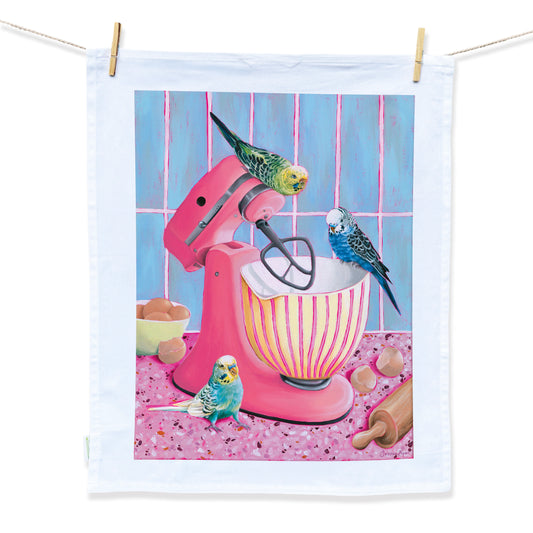 Australian Artist Jessica Alpert designed and hand painted the illustration of three blue and green budgie birds around a pink kitchen stand mixer with a pink and yellow striped mixing bowl in a colourful kitchen. the illustration is placed on a white tea towel with a white border and is hanging on a string attached by two wooden pegs.