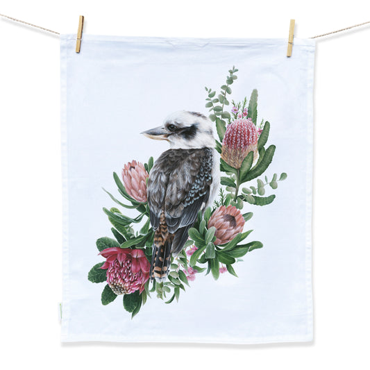 A white tea towel with a design hand painted by Australian Artist Jessica Alpert, of an Australian Kookaburra bird looking to his left peacefully, perched amongst pretty green and pink native foliage and flowers, against a soft baby blue background.