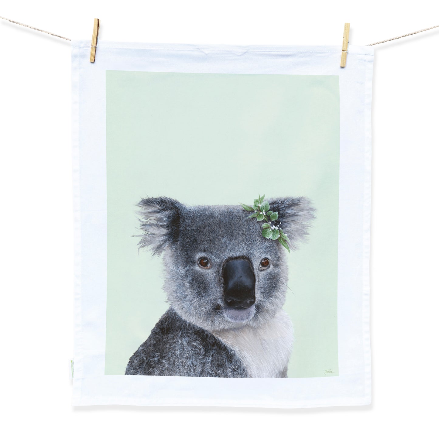 Australian Artist Jessica Alpert designed and hand painted the fluffy koala illustration. The koala is on a pale green background and has a leaf flower crown on it's right ear. The illustration is on a white tea towel with a white border hanging from two wooden pegs on a string line.