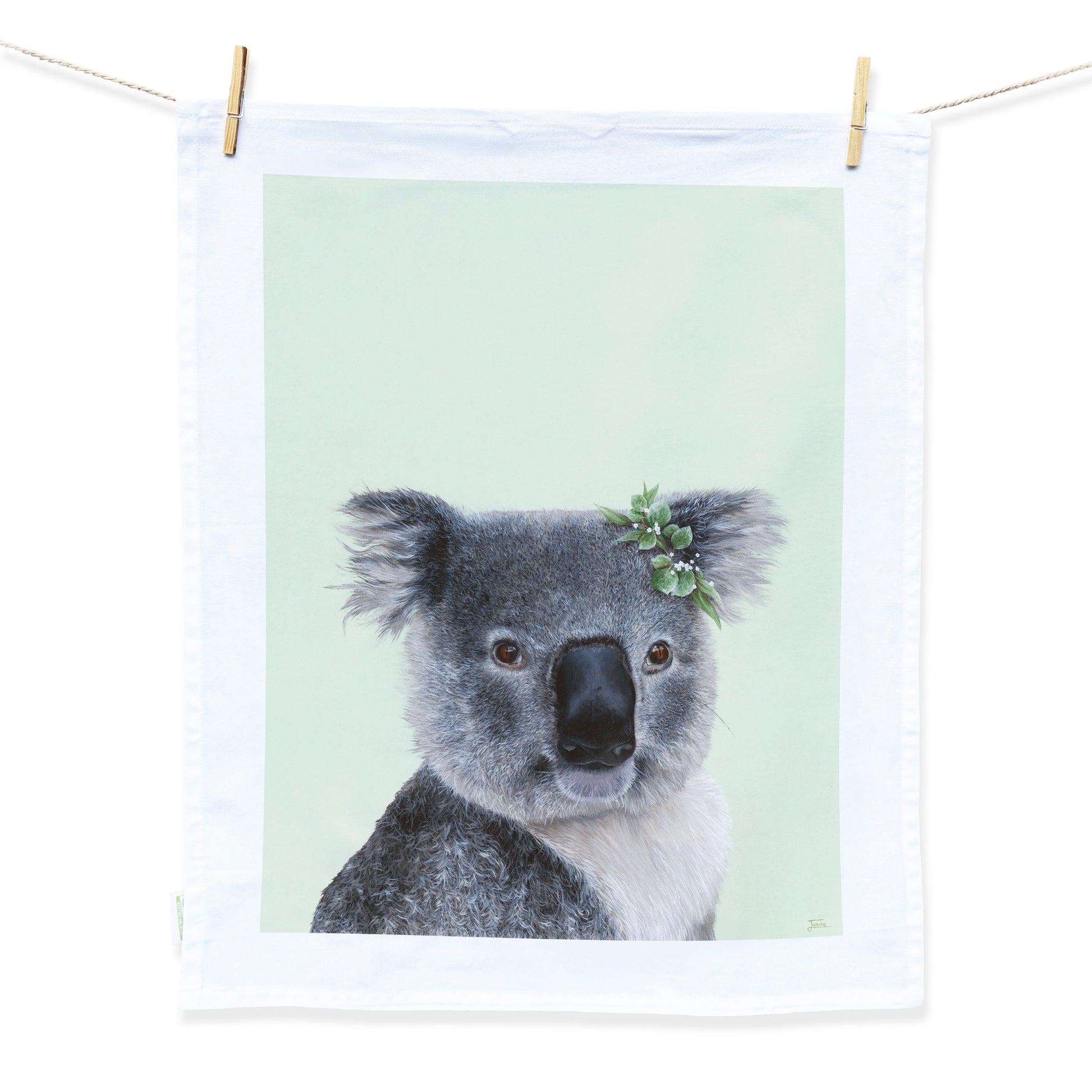 Australian Artist Jessica Alpert designed and hand painted the fluffy koala illustration. The koala is on a pale green background and has a leaf flower crown on it's right ear. The illustration is on a white tea towel with a white border hanging from two wooden pegs on a string line.