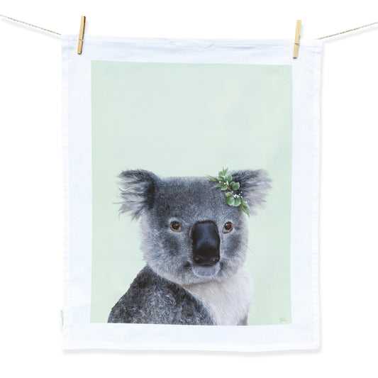Australian Artist Jessica Alpert designed and hand painted the fluffy koala illustration. The koala is on a pale green background and has a leaf flower crown on it's right ear. The illustration is on a white tea towel with a white border hanging from two wooden pegs on a string line.